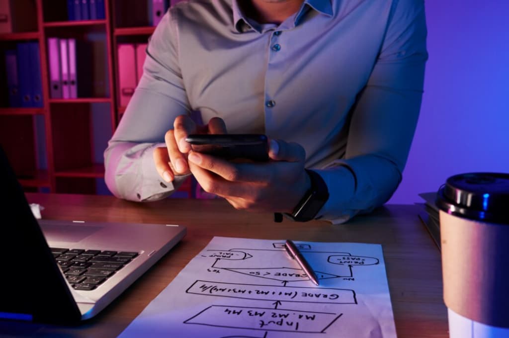 A person using phone at a desk lit by a computer screen, with UX design sketches on the table